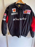 Front view of a black Ferrari Formula 1 men’s jacket with red accents on sleeves and multiple sponsor logos including Shell, Pirelli, Santander, Ferrari on chest and arms.