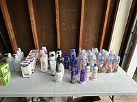 Wide view of assorted personal care products including white and purple bottles, boxed liquid soaps, blue and purple hair care products, and pump bottles arranged on a folding table.