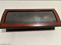 Closed rectangular wooden watch case with warm brown finish and clear glass top display panel, sitting on a white surface.