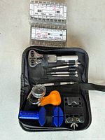 Photo showing various watch repair tools inside a black zippered case including screwdrivers, a case opener, tweezers, loupe, orange tool, blue holder, and black strap holder.