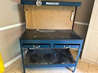 Front view showing work surface, two drawers, lower shelf, and pegboard back with hooks.