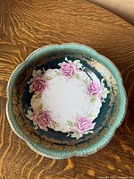 Top view of antique Austrian porcelain plate showing hand-painted pink roses on white center, dark teal background, gold decorative border, and scalloped textured teal rim.