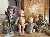 Four antique dolls standing in front of a wooden backdrop. The dolls include a porcelain doll with checked dress, a naked composition doll, a baby doll in beige outfit, and a larger doll in patterned clothes.
