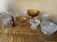 Seven antique glass pieces arranged on a wooden surface including clear and amber colored bowls with Moon and Stars pattern. A small red glass lantern is placed among them.