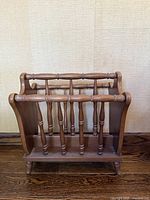 Front and side view of antique wooden magazine holder showing spindle bars and curved handles, placed on wood floor against textured beige wall.