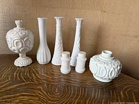 Full set of six white milk glass items arranged side by side on wooden surface against burlap background, showing variety of shapes and textures.