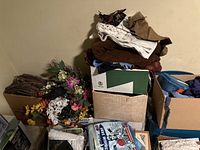 Wide view showing stacked bins and boxes containing assorted fabric, yarn, and artificial flowers on the floor against a wall.