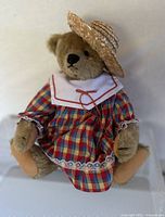 Front view of the Margaret Strong Steiff collectible bear dressed in a plaid dress and straw hat sitting against a plain background.