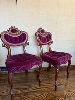 Pair of Victorian antique armchairs with burgundy velvet upholstery and carved wooden frames shown side by side on wooden floor against a light wall.