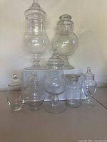 Front view showing seven clear glass vintage apothecary jars with different shapes and sizes placed on a flat surface in front of a white background.