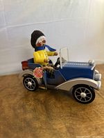 Side angled photo showing the blue antique-style car with a driver figurine dressed in yellow and black hat, blue scarf and plaid pants. The detailed car includes black and white wheels, side storage box, and windshield.