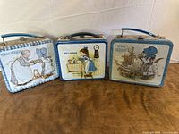 Three vintage Holly Hobbie metal lunch boxes with blue handles lined side by side on a wood surface against a white wall.