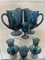 Front view of two blue carnival glass pitchers and five various footed glasses, all featuring Harvest Grape grape and leaf pattern with iridescent sheen.