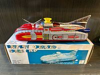Front and side view of the red and silver tin space ship toy sitting on its original box showing colorful branding and ship image.