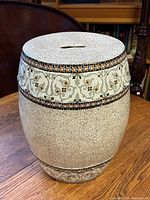 Side angle photo showing ceramic garden stool with speckled finish and mosaic floral-like band around upper circumference.