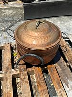 Copper pot with lid on, showing hammered texture and metal handles, sitting on wooden pallet outside