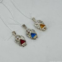 Three sterling silver angel charms with red, blue, and yellow/orange triangular colored gemstones and round clear stones, laid out on a white background.