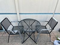 Full set of outdoor furniture: one round metal grated table and two matching metal grated chairs with armrests, set on patio surface.
