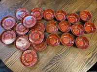 Elevated view showing 11 red-orange lacquered Japanese bowls and plates arranged in rows on a wooden surface, featuring black and green floral designs.