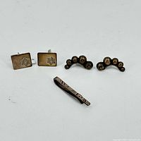 Sterling silver rectangular cufflinks with star design, silver tie clip with linear engraving, and spherical graduated silver earrings arranged on white background