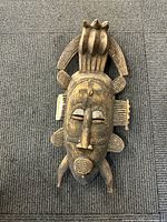 Front view of wooden tribal mask placed on carpeted floor showing full mask design including facial features and top crest.