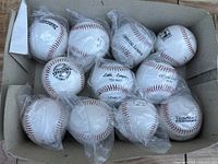 Photo showing 11 baseballs in a box, some individually wrapped in plastic. Different sizes and types visible including Official League and Little League Tee Ball.
