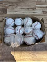 Top-down view of 11 white tee balls with red stitching inside a cardboard box. Some balls have logos visible including Easton and Wilson.