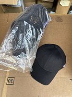 Black MLB team cap still in original plastic packaging beside an unpackaged identical cap showing front and side MLB logo.