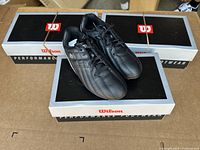 Three pairs of black and silver Wilson youth soccer cleats arranged on Wilson shoeboxes, showing side and top views.