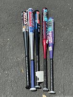 Five aluminum baseball bats lying on asphalt, showing full length of bats and wrapped handles.
