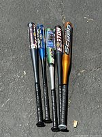 Full view of five aluminum baseball bats laid out on pavement showing black grips and various colors and brands.