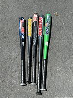 Five aluminum baseball bats laid on pavement, showing barrels and handle grips clearly, different colors and brands visible