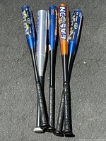 Group of 5 aluminum baseball bats lying on pavement, showing full length and colors including blue, silver, black, and orange.