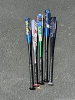 Group of 5 aluminum baseball bats laid on asphalt, showing full length from handle grips to barrels, colors include blue, silver, green, and dark.