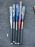 Group of 5 aluminum baseball bats laid on asphalt, showing grips and barrel logos.