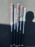 Five aluminum bats laid out side by side on pavement showing length and condition, with black grips and silver barrels.