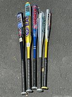 Five aluminum baseball bats lying side by side on asphalt, showing full length and branding on barrels.