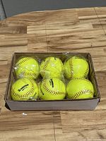 Six yellow Rawlings softballs in a cardboard box, wrapped in plastic, placed on a wooden surface.