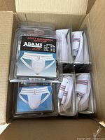 Photo of all 9 Adams adult supporter cups in original packaging inside a cardboard box, showing jockstraps with cups enclosed, some with red and blue detail bands.