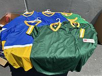 Six Wilson sports tops displayed flat with hangers: three green with gold trim, and three blue and gold.