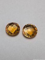 Two round faceted amber stones placed on a plain background. Both display a golden-brown color and checkered faceting that reflects light.