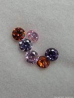 Photo showing six assorted round cut genuine gemstones in purple, pink, and orange hues arranged in a curve on a white background.
