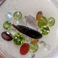 Top-down view of 18 assorted faceted genuine gemstones on a white background including green peridots, dark garnet, red carnelian, clear quartz/moonstone, purple amethyst, and small blue stones.