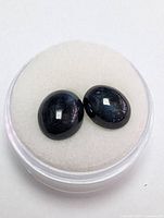 Photo showing pair of dark blue oval cabochon sapphire stones inside round container with white padded cushion.