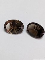 Two faceted oval cut genuine smokey quartz gemstones viewed from different angles showing reflective facets and transparent brown-gray color.