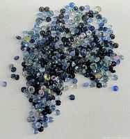 Close-up of numerous small sapphire gemstones scattered on a white surface showing variety of colors including blue, green, yellow, purple and black with mixed cuts.
