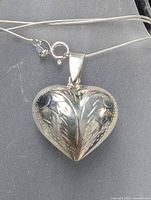 Close-up image of silver heart pendant with floral embossing on a silver chain, showing pendant details and chain clasp.