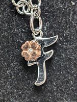 Close-up of silver bracelet charm showing stylized letter 'F' with gold-tone flower and clear center stone.