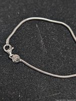 Close-up photo showing the lobster clasp and part of the silver snake chain bracelet on a black background.