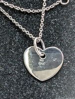 Close-up photo of the heart-shaped silver pendant with chain on a black fabric background showing 925 stamp and brand Etui mark on the pendant.